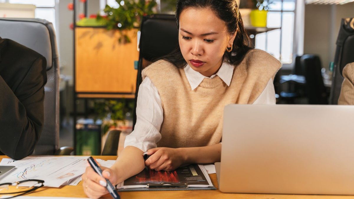 Karyawati kantor mencatat data di samping laptop dan lembar grafik di meja kerja