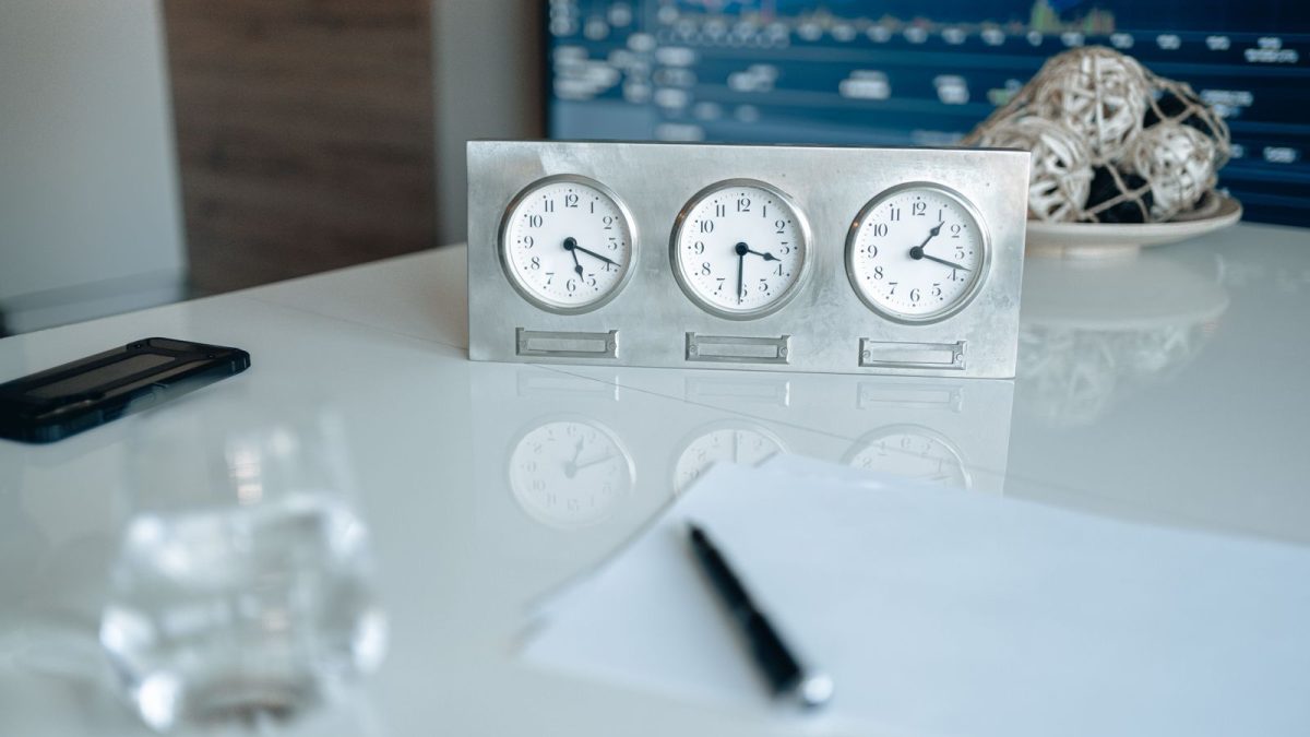 Tiga jam analog berjajar di atas meja kantor putih, dengan kertas dan pulpen di depannya