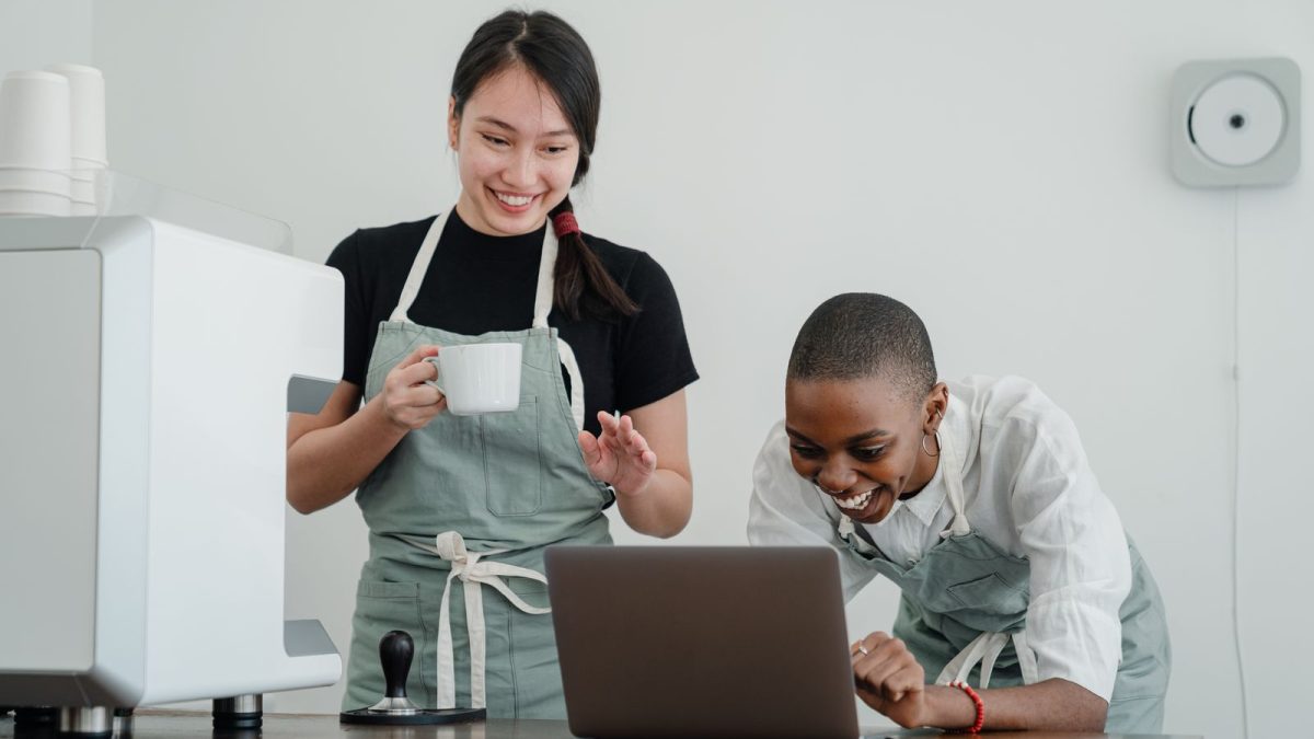Barista senior menjelaskan langkah kerja di laptop kepada rekan baru di dapur kafe
