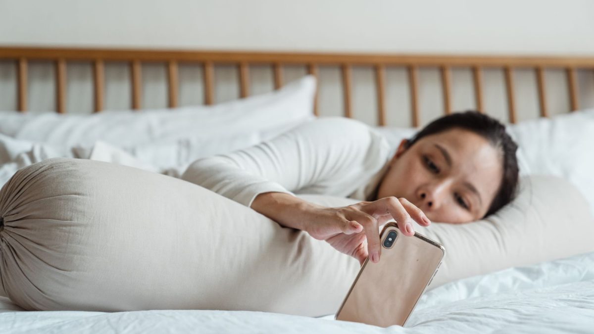 Perempuan berbaring di kasur sambil meraih ponsel di dekat bantal guling