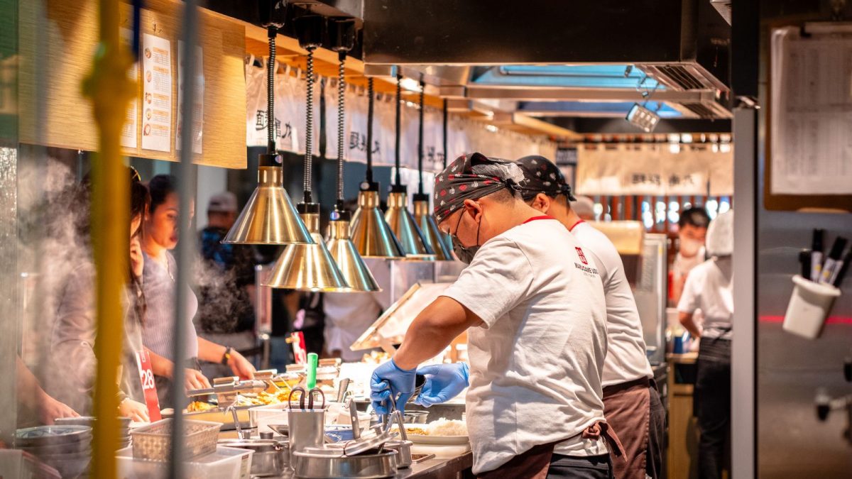 Juru masak bersarung tangan biru menyiapkan pesanan di dapur restoran yang sibuk bersama rekan satu shift