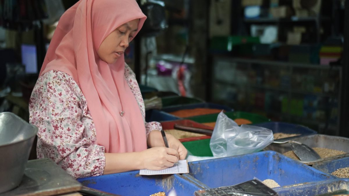 Pedagang perempuan berhijab mencatat penjualan di samping kalkulator pada lapak pasar