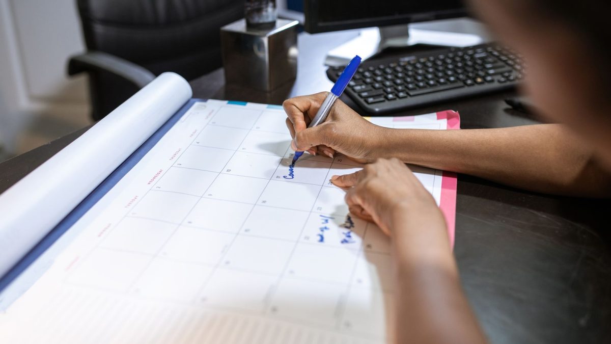 Tangan karyawan menulis jadwal pada kalender meja berukuran besar di samping keyboard kantor