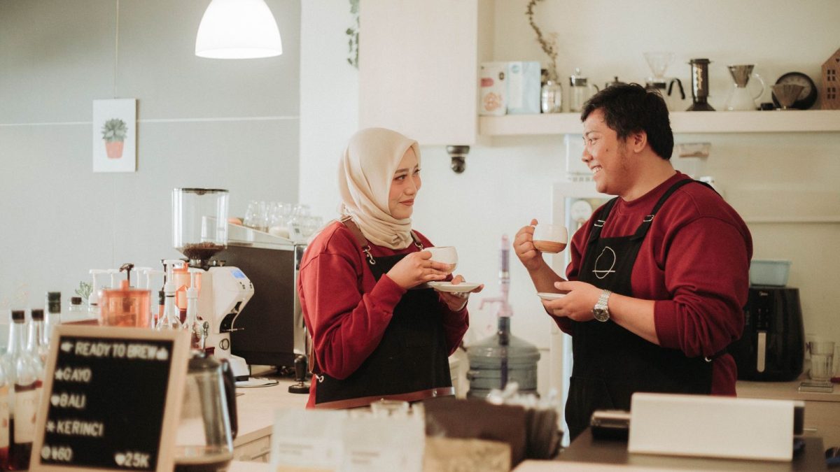 Dua barista berbincang santai sambil memegang cangkir kopi di balik meja bar kafe