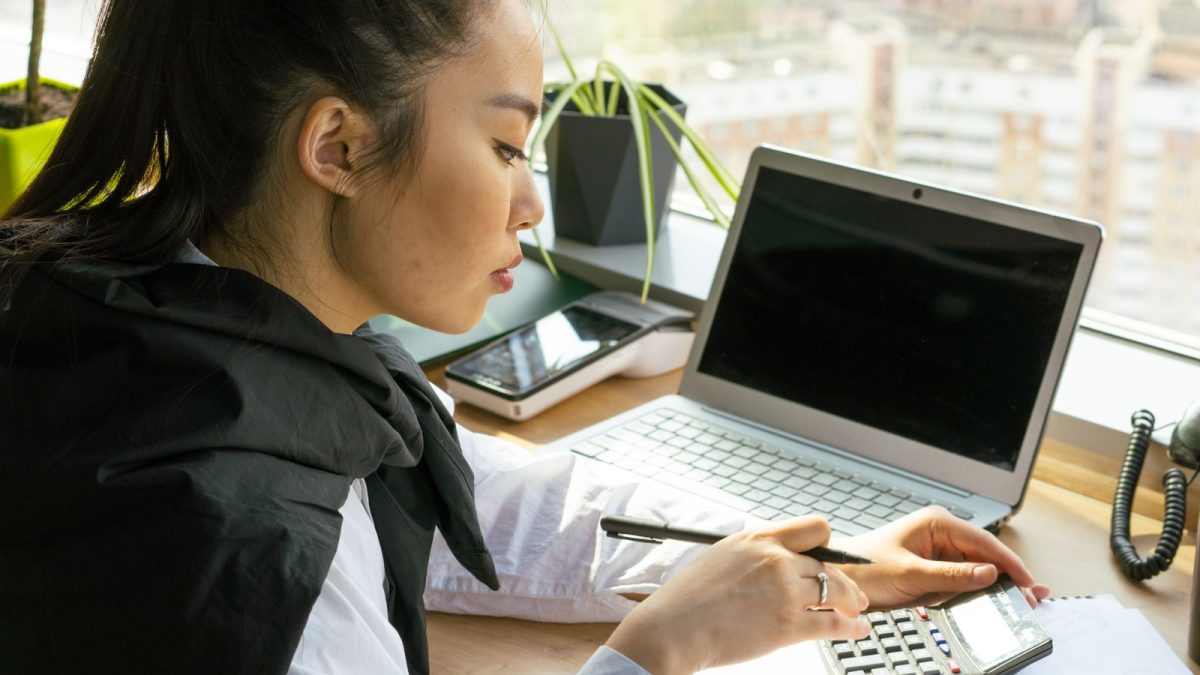 Karyawati menghitung anggaran dengan kalkulator di samping laptop di meja dekat jendela