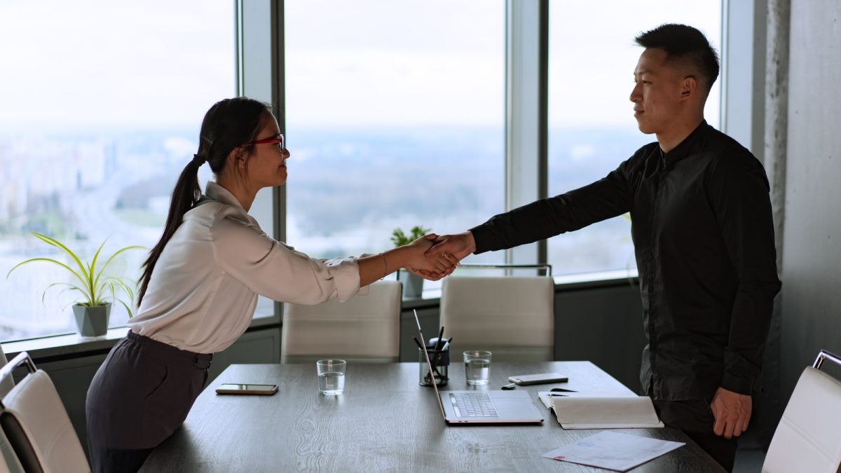 Perempuan berkacamata dan pria berkemeja hitam berjabat tangan di ruang rapat kantor berjendela kaca besar